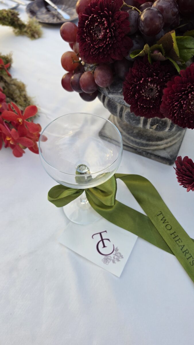 Taça de espumante com fita de cetim verde personalizada, guardanapo com monograma e dálias bordeaux com uvas